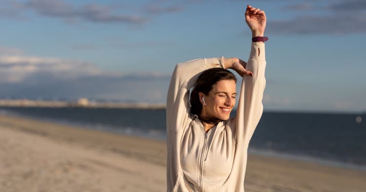 Μια νεαρή γυναίκα με κλειστά μάτια και ένα πλατύ χαμόγελο τεντώνει τα χέρια της πάνω από το κεφάλι της στην αμμουδιά. Φοράει μπεζ ζακέτα με κουκούλα, ασύρματα ακουστικά και ένα αθλητικό ρολόι, με φόντο την ήρεμη θάλασσα και τον γαλάζιο ουρανό.